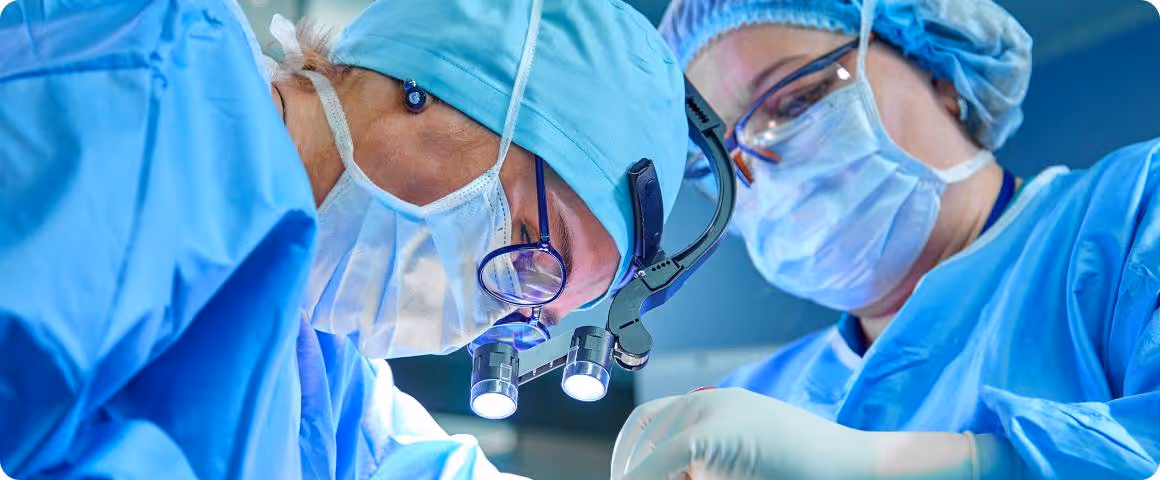 Hai bác sĩ mặc đồ mổ màu xanh tập trung thực hiện thủ thuật, một người sử dụng kính lúp phẫu thuật có đèn.